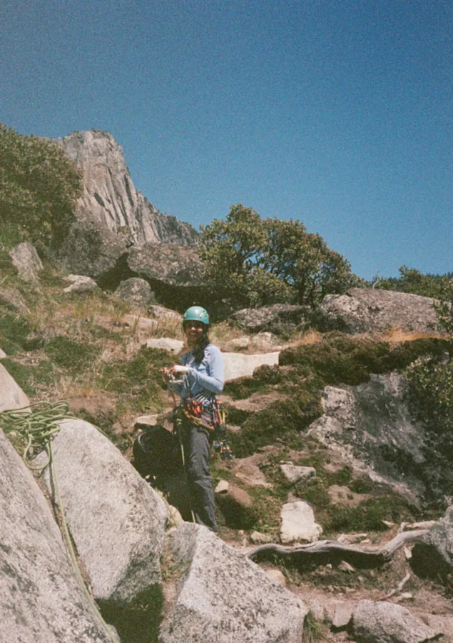 Nitya in a climbing helmet and harness at the base of a granite crag